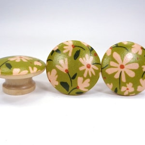 May include: Three wooden knobs with a floral pattern. The knobs are green with pink flowers and have a natural wood base.