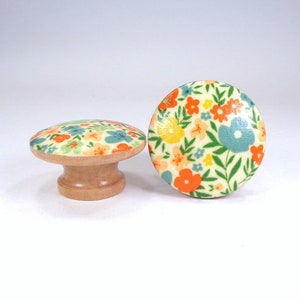 May include: Two round wooden cabinet knobs featuring a flat top surface decorated with a colorful floral pattern. The design includes small orange, yellow, and light blue flowers with green leaves on a cream background. The base is natural wood.