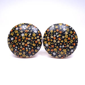 May include: Two round, floral-patterned earrings. The earrings have a black background with small orange, yellow, and white flowers. The earrings are set against a white background.