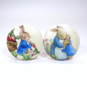 May include: Two round cabinet knobs with illustrations of Peter Rabbit. One knob shows Peter Rabbit with a bag of carrots, the other shows a rabbit embracing another rabbit. The knobs have a cream-colored background.