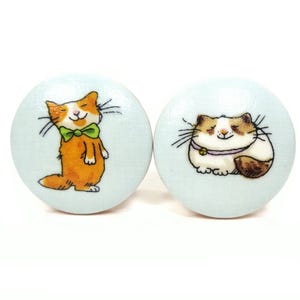 May include: Two round, light blue decorative knobs. One knob features an orange cat wearing a green bow tie. The other knob has a brown and white cat with a purple collar. Both cats have black whiskers and are smiling.