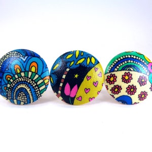 May include: Three round, colorful, wooden buttons with different patterned designs. The buttons are blue, yellow, and green with various shapes and colors.