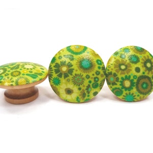 May include: Three decorative wooden knobs with a floral pattern in shades of green, yellow, and teal. One knob is mushroom-shaped, while the other two are round. The knobs are set against a white background.