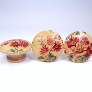 May include: Three wooden knobs with a floral design. The knobs are round and have a beige background with red and green flowers.