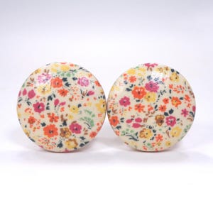 May include: Two circular buttons featuring a cream-colored base with a dense, multi-colored floral pattern. The print includes small flowers in shades of orange, pink, yellow, and green, distributed evenly across the smooth, rounded surface of each button.