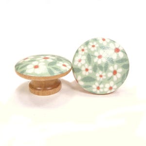 May include: Two decorative wooden cabinet knobs. Each knob features a floral design with white flowers, red centers, and green leaves on a light green background. The knobs have a natural wood base.