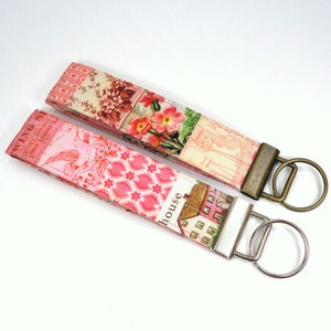 May include: Two pink and white fabric keychains with floral and house patterns. One keychain has a bronze key ring and the other has a silver key ring.