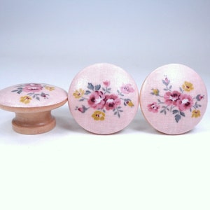 May include: Three pink wooden knobs with a floral design. The knobs have a light pink background with a floral pattern in shades of pink, yellow, and gray.