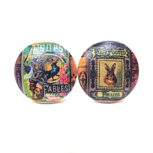 May include: Two round, colorful, wooden buttons with vintage-style illustrations. One button features a peacock, a bird, and the text "Aesop's Fables". The other button features a rabbit and the text "Four Footed Friends".