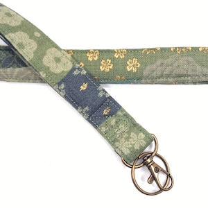 May include: A green and blue patterned lanyard with a gold-colored clasp and key ring. The lanyard features a floral design with gold accents. The fabric is a patchwork style with various patterns and colors.