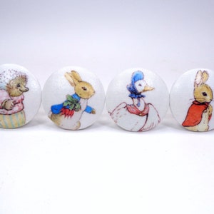 May include: Four white, round cabinet knobs featuring illustrations of characters from Beatrix Potter's stories. The knobs depict a hedgehog, two rabbits, and a duck, each in colorful attire. These knobs are perfect for a child's room or a whimsical touch.