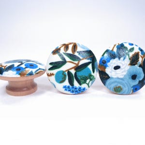 May include: Three decorative cabinet knobs with floral patterns. The knobs feature a white background with blue, teal, and brown floral designs. One knob is a small, round table-like shape with a wooden base. The other two are round.
