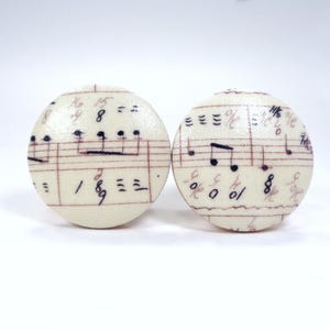 May include: Two circular knobs featuring a cream-colored fabric surface printed with black musical notes and red staff lines. The design displays various musical symbols, including clefs, notes, and time signatures, arranged across the round faces.