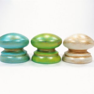 May include: Three decorative wooden knobs in a row, each with a unique color. The knobs are painted in teal, green, and off-white with gold accents. The knobs have a mushroom-like shape and are set against a white background.