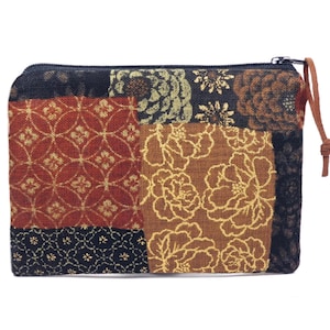 May include: A rectangular zippered pouch featuring a patchwork design of various fabric panels. The patterns include black, rust red, and brown sections with gold-colored floral and geometric motifs. A thin brown leather cord is attached to the zipper pull.