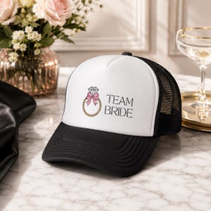 May include: A black and white trucker hat featuring a graphic of a gold ring with a diamond and a pink bow, accompanied by the text "TEAM BRIDE" in black lettering. The cap has a solid white front panel, a black brim, and a black mesh back.