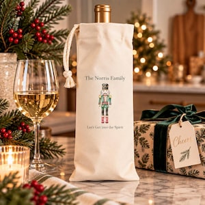 Könnte beinhalten: Ein cremefarbener Weinflaschen-Geschenkbeutel aus Stoff mit Kordelzug. Der Beutel ist mit einer Illustration eines Nussknacker-Soldaten sowie dem Text "The Norris Family" und dem Schriftzug "Let's Get into the Spirit" in Schwarz bedruckt.