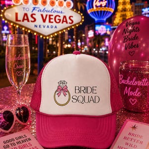May include: A bright pink and white trucker hat featuring a graphic of a gold diamond ring with a pink bow and the text "BRIDE SQUAD". The hat is surrounded by a champagne flute, a pink balloon, heart-shaped sunglasses, and two cards.