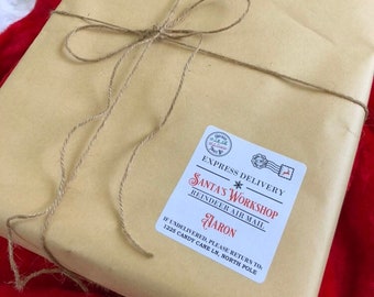 May include: A wrapped package in tan paper tied with twine, featuring a label that reads "Santa's Workshop, Reindeer Air Mail" and the name "Aaron." The package rests on a red and white surface, evoking a festive holiday theme.
