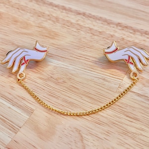 Puede incluir: Dos broches de mano de esmalte blanco, unidos por una cadena dorada. Cada broche presenta detalles rojos y contornos negros y dorados. Los broches se presentan sobre una superficie de madera clara.