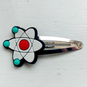 May include: A white and black atom-shaped hair clip with a red centre and turquoise accents. The clip is made of plastic and has a silver metal bar.