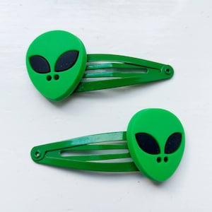 May include: A pair of green hair slides, each with a green alien head and black eyes. The slides are made of metal and are a bright green colour. The alien heads are a lighter shade of green.