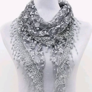 Puede incluir: Una bufanda de encaje gris con bordados florales y detalles de flecos. La bufanda está drapeada sobre un maniquí, mostrando su delicado diseño y textura. Los intrincados patrones y el color suave la convierten en un accesorio versátil.