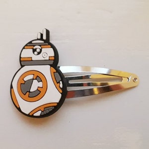 Puede incluir: Una pinza para el pelo de metal plateado con un diseño del personaje BB-8. El diseño de BB-8 es blanco, naranja, gris y negro. La pinza para el pelo es de estilo horquilla, adecuada para sujetar el cabello.