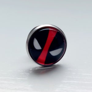 Puede incluir: Un pin de plata con un logotipo de Deadpool rojo y negro. El logotipo es una cara estilizada con una barra roja sobre los ojos.