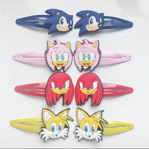 Könnte beinhalten: Eine Sammlung farbenfroher Haarspangen mit Charakteren aus der Sonic the Hedgehog-Franchise. Die Spangen zeigen Sonic, Amy Rose, Knuckles und Tails, jeweils mit ihren charakteristischen Farben und Designs. Auf weißem Hintergrund.