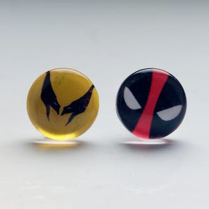 Può includere: Un paio di orecchini rotondi e colorati con loghi di supereroi. L'orecchino sinistro è giallo con un logo Wolverine nero. L'orecchino destro è nero con un logo Deadpool rosso.