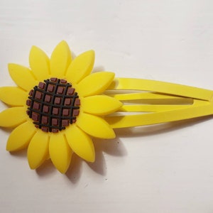 以下が含まれることがあります： ひまわり柄の黄色いプラスチック製のヘアクリップ。ひまわりは、格子模様の茶色の芯を持っています。