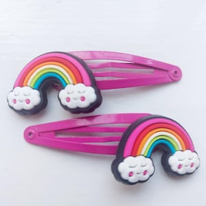 Op de afbeelding: Twee roze haarspeldjes met regenboog- en wolkenontwerpen. Elk speldje heeft een kleurrijke regenboog met rode, oranje, gele, groene en blauwe bogen, en witte wolken met lachende gezichten.