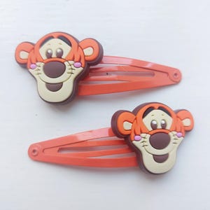 以下が含まれることがあります： オレンジ色のヘアクリップ2つ。それぞれに漫画のトラの顔が描かれています。トラはオレンジと茶色の毛皮、ベージュの口吻、ピンクの頬をしています。クリップはオレンジ色の金属製で、髪をまとめるように設計されています。
