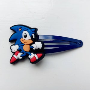 Könnte beinhalten: Blauer und roter Plastik-Haarklipp mit einem Cartoon-Bild von Sonic the Hedgehog, der rennt. Der Clip ist dunkelblau und hat einen Federmechanismus.