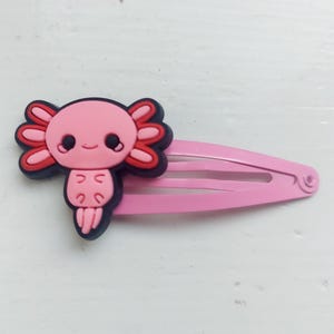 Może przedstawiać: Różowa plastikowa spinka do włosów z kreskówkowym wzorem aksolotla. Axolotl jest różowy z czerwonymi skrzelami i czarnymi oczami.