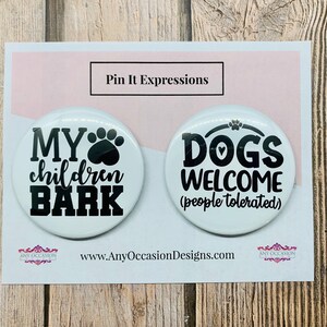 May include: Two white round button pins with black text. The first pin says "MY children BARK" with a paw print and heart. The second pin says "DOGS WELCOME (people tolerated)" with a paw print and heart.