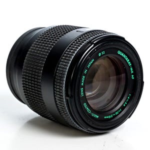 May include: Black Quantaray-MX AF camera lens with a textured grip. The lens has a diameter of 52mm and a focal length of f=70-210mm. The text "LENS MADE IN JAPAN" and "MULTI-COATED" are visible. The lens is angled on a white surface.