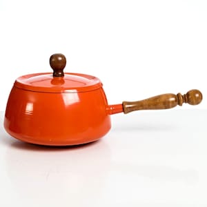 May include: A vibrant orange saucepan with a matching lid and a wooden handle. The handle is attached to the side of the pan. The lid has a dark brown knob. The pan is on a white surface.