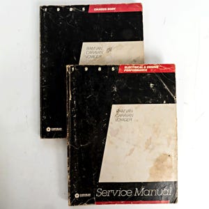 May include: Two 1985 Chrysler Corporation service manuals for Ram Van, Caravan, and Voyager vehicles. The black and white covers feature red accents, with one manual labeled Chassis/Body and the other labeled Electrical & Engine Performance.