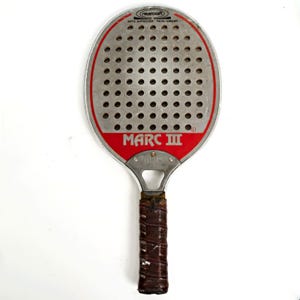 May include: A Marc III platform tennis paddle with a silver perforated face, a red border, and a brown wrapped handle. The paddle features the text Marcraft, APTA Approved, and Marc III printed on the face, with a metal throat and a small cutout.