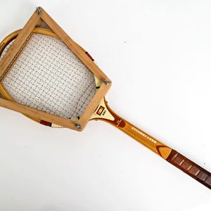 May include: A vintage wooden tennis racket secured within a wooden press frame. The racket features a light brown wood finish, a white string pattern, and a brown leather grip. The text 440 and MASTERBUILT are visible on the racket throat.