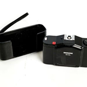May include: A black Minox 35 EL camera with its matching black leather case. The camera has a silver flash mount and a red button. The case is open, revealing its interior. The camera is labeled "MINOX 35 EL".