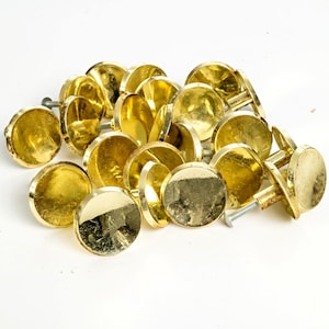 May include: A collection of shiny, gold-colored cabinet knobs. Each knob has a round, flat face and a screw for attachment. The knobs are scattered on a white surface, reflecting light and creating highlights.