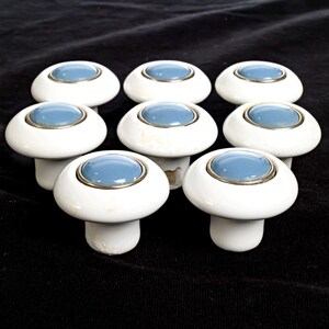 May include: A set of eight round cabinet knobs featuring a smooth white ceramic base and a central circular blue inlay surrounded by a thin silver-toned metal rim. Each knob has a mushroom-shaped profile with a cylindrical stem for mounting.