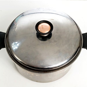May include: A round stainless steel cooking pot with a matching lid. The pot features two black side handles and a central lid knob with a black rim and a light peach-colored center. The text "Duncan Hines" is printed on the lid knob.