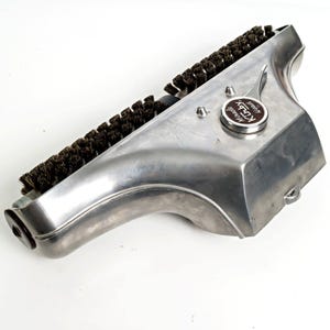 May include: A vintage Kirby vacuum cleaner attachment. The attachment is made of brushed aluminum and features a row of dark bristles. A circular emblem reads "Kirby Miracle Wood".