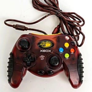 May include: A translucent dark red wired game controller for Xbox featuring a Mad Catz logo, black rubberized side grips, a black directional pad, two black analog sticks, and four colored action buttons labeled A, B, X, and Y.