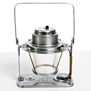 May include: A vintage chrome-plated metal lantern featuring a clear glass cylindrical shade, a ribbed top housing with a black switch, and a sturdy rectangular base. A tall, arched metal handle is attached to the sides for carrying.