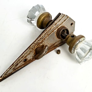 May include: A vintage door hardware set featuring two clear, faceted glass knobs attached to a tarnished, triangular metal backplate. The metal components show visible oxidation and rust, with a small metal screw resting near the central spindle.